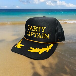 May include: Black trucker hat with a yellow embroidered design. The hat says "Party Captain" in yellow lettering.