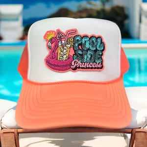 May include: A trucker hat with a white front panel and a coral-colored brim and mesh back. The front features a patch with a pink flamingo float, a cocktail, and the words "Pool Side Princess" in sequined letters.