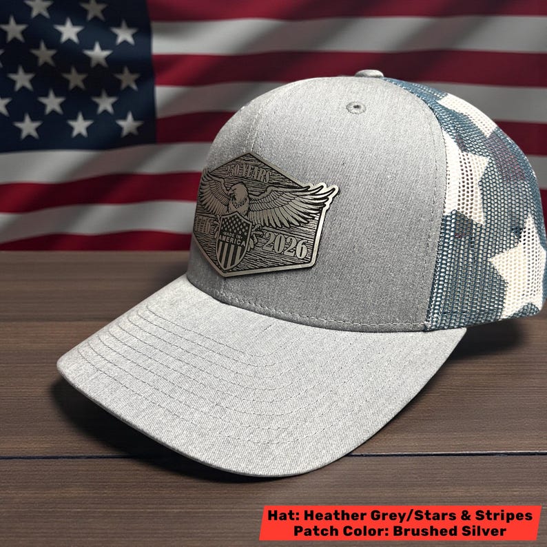 May include: Heather grey baseball cap with a mesh back featuring a star pattern. The front displays a brushed silver patch with an eagle, shield, and the text "2026". The hat's brim is solid grey.