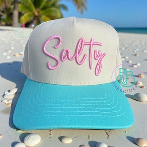 May include: A light gray and turquoise baseball cap with the word "Salty" embroidered in pink script across the front. The hat has a flat brim and a structured crown, perfect for beach or casual wear.