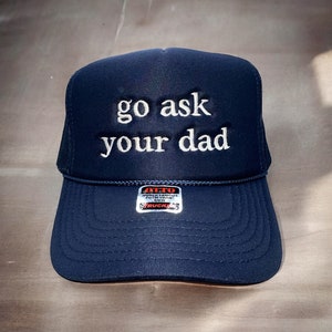 May include: Navy blue trucker hat with the embroidered text "go ask your dad" in white. The hat features a mesh back and a curved brim. A small tag is visible on the front.