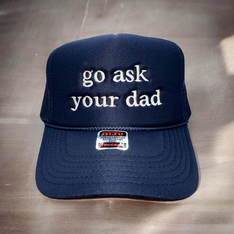 May include: Navy blue trucker hat with the embroidered text "go ask your dad" in white. The hat features a mesh back and a curved brim. A small tag is visible on the front.