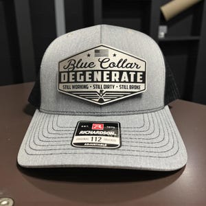 Peut inclure: Casquette de camionneur grise avec un dos en filet noir et une visière incurvée. L'avant présente un écusson avec les mots "Blue Collar DEGENERATE" et les phrases "STILL WORKING STILL DIRTY STILL BROKE". Avec une étiquette Richardson.