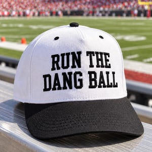 May include: A white baseball cap with a black brim and a black button on top. The front of the cap features the embroidered black text "RUN THE DANG BALL" in a bold, block-style font. The cap has ventilation eyelets on the crown panels.