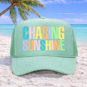 May include: A light green trucker hat with the words "CHASING SUNSHINE" in colorful block letters. The hat has a mesh back and a curved brim. The background shows a beach with a palm tree and blue ocean.