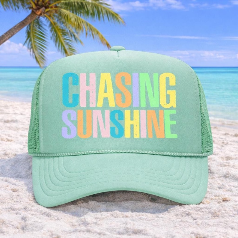 May include: A light green trucker hat with the words "CHASING SUNSHINE" in colorful block letters. The hat has a mesh back and a curved brim. The background shows a beach with a palm tree and blue ocean.