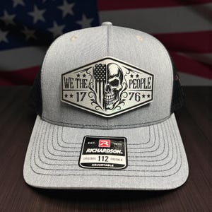 May include: Gray trucker hat with a black mesh back and a patch featuring a skull, American flag, and the text "WE THE PEOPLE 1776". The hat has a curved brim and an adjustable strap.