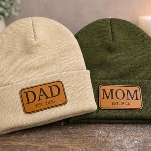 May include: Two knitted beanies, one beige and one olive green, with leather patches. The beige beanie has "DAD EST. 2026" and the green beanie has "MOM EST. 2026". The beanies are on a wooden surface.