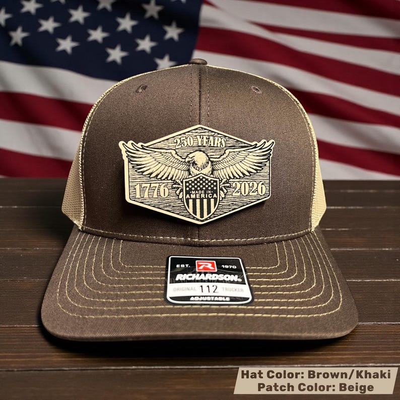 May include: A brown and khaki trucker hat with a beige patch featuring an eagle, shield, and the text "250 YEARS 1776-2026 AMERICA". The hat has a curved brim and adjustable strap. The Richardson brand label is visible.