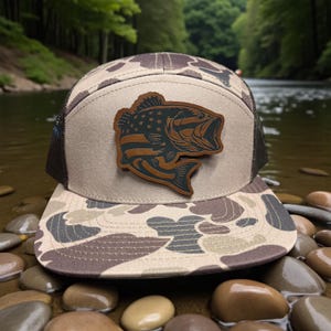 May include: A camouflage-patterned trucker hat with a brown leather patch featuring a fish design with an American flag motif. The hat has a mesh back and a flat brim. The hat is sitting on a bed of rocks near a river.