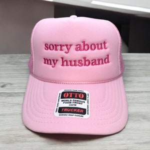 May include: Pink trucker hat with a mesh back and the text "sorry about my husband" embroidered in pink on the front. The hat has a black label with the text "OTTO WORLD FAMOUS FOAM FRONT OSFM TRUCKER DURABLE SNAP CLOSURE" on it.