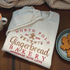 May include: Beige sweatshirt with embroidered text "North Pole Gingerbread Bakery" in red and gold. Features a gingerbread man and festive icons. A plate of gingerbread cookies is on the right.
