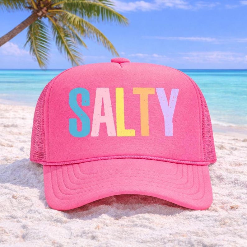 May include: A bright pink trucker hat with the word "SALTY" in colorful letters across the front. The hat has a mesh back and a curved brim. The hat is sitting on a sandy beach with a palm tree and ocean in the background.