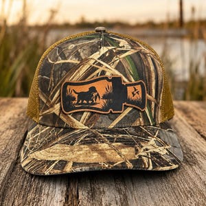 May include: A camouflage print trucker hat featuring a brown mesh back and a brown leather patch on the front. The patch is shaped like a duck call and displays a silhouette of a dog retrieving a bird in a marsh with flying ducks.