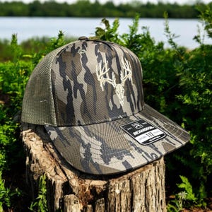 May include: Camouflage pattern baseball cap with a white embroidered deer skull design. The cap has a mesh back and a curved brim. The Richardson brand label is visible on the brim. The cap is sitting on a tree stump.