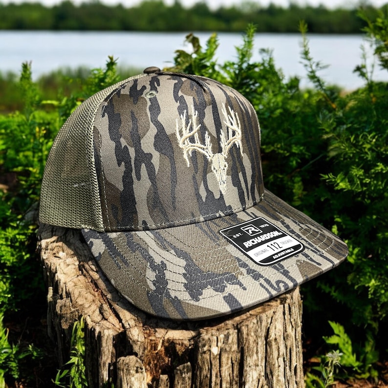 May include: Camouflage pattern baseball cap with a white embroidered deer skull design. The cap has a mesh back and a curved brim. The Richardson brand label is visible on the brim. The cap is sitting on a tree stump.