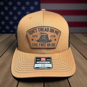 Peut inclure: Casquette de camionneur beige et noire avec un écusson portant l'inscription "DON'T TREAD ON ME" avec un serpent enroulé. La casquette porte également l'inscription "LIVE FREE OR DIE" et "ESTD. 1776". La casquette porte une étiquette Richardson indiquant "ORIGINAL 112 TRUCKER ADJUSTABLE".