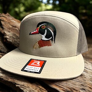 May include: A beige and grey flat bill snapback hat with an embroidered wood duck design. The hat has a mesh back and a red label that says "Remarkable Flat Bill Snapback Est. 1999".