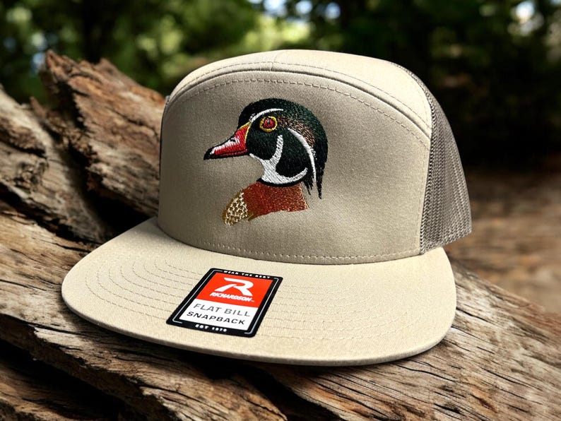 May include: A beige and grey flat bill snapback hat with an embroidered wood duck design. The hat has a mesh back and a red label that says "Remarkable Flat Bill Snapback Est. 1999".