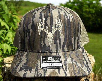 May include: A camouflage trucker hat featuring a white embroidered deer skull and antler design on the front panel. The cap has a mesh back, a structured crown, and a sticker on the brim that reads Richardson 112 Original Adjustable.