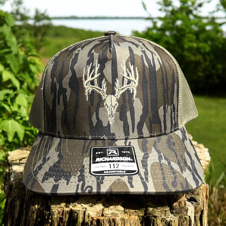 May include: Camouflage trucker hat with a deer skull and antler design. The hat features a curved brim, mesh back, and an adjustable strap. The Richardson brand label is visible on the front.
