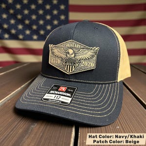 May include: Navy and khaki trucker hat with a beige patch. The patch features an eagle, shield, and the text "250 YEARS 1776 AMERICA 2026". The hat has a curved brim and an adjustable strap. The brand "RICHARDSON" is visible.