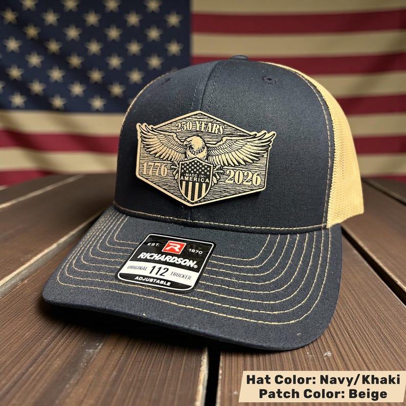 May include: Navy and khaki trucker hat with a beige patch. The patch features an eagle, shield, and the text "250 YEARS 1776 AMERICA 2026". The hat has a curved brim and an adjustable strap. The brand "RICHARDSON" is visible.