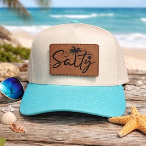 May include: A cream-coloured trucker hat with a bright turquoise brim and a rectangular brown leather patch on the front. The patch features the word Salty in black script, accompanied by a small palm tree and sun graphic design.