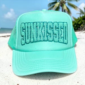 May include: A turquoise trucker hat with the word "SUNKISSED" embroidered in sequins across the front. The hat features a curved brim and a mesh back, perfect for sunny days. The hat is sitting on a sandy surface.