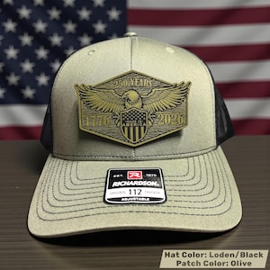 May include: Olive green and black trucker hat with a gold patch featuring an eagle, the years 1776 and 2026, and the text "250 YEARS" and "MADE IN AMERICA". The hat is adjustable and has a Richardson 112 Trucker label.