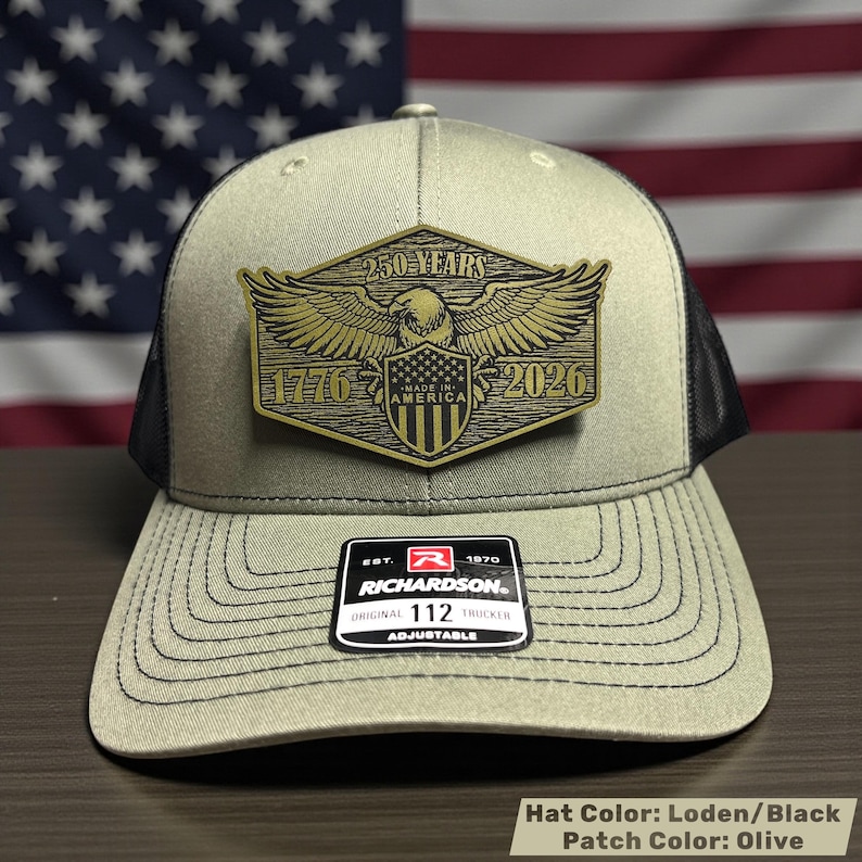May include: Olive green and black trucker hat with a gold patch featuring an eagle, the years 1776 and 2026, and the text "250 YEARS" and "MADE IN AMERICA". The hat is adjustable and has a Richardson 112 Trucker label.