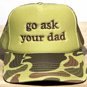 May include: A green trucker hat with the phrase "go ask your dad" embroidered in brown. The hat features a camouflage pattern on the brim and a mesh back.