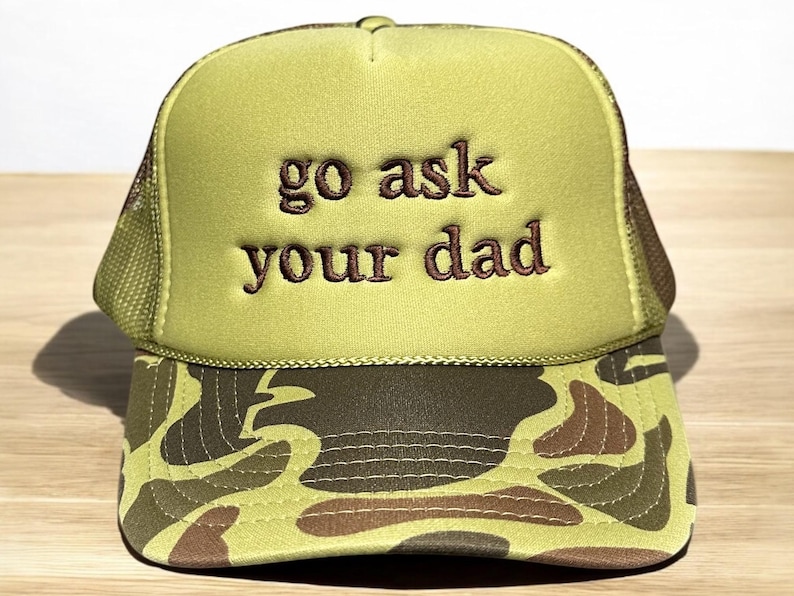 May include: A green trucker hat with the phrase "go ask your dad" embroidered in brown. The hat features a camouflage pattern on the brim and a mesh back.