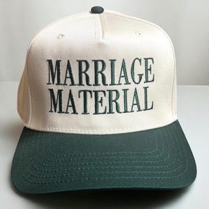 May include: A white baseball cap with a green brim. The front of the cap has the text "MARRIAGE MATERIAL" embroidered in green.