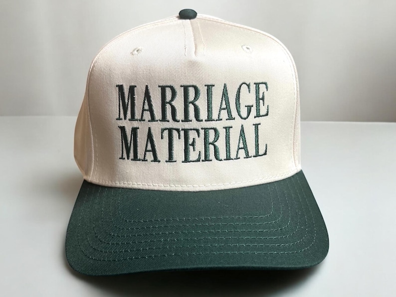 May include: A white baseball cap with a green brim. The front of the cap has the text "MARRIAGE MATERIAL" embroidered in green.