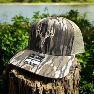 May include: Camouflage baseball cap with a deer skull and antler design. The cap has a mesh back and a curved brim. The front of the cap features a white embroidered deer skull and antler design. The tag reads "RICHARDSON 112".