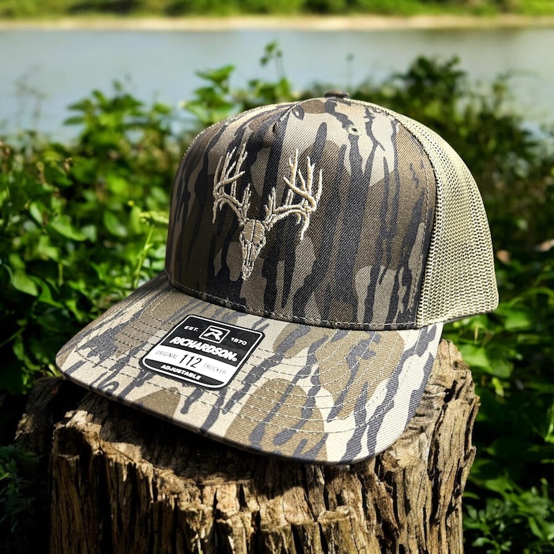 May include: Camouflage baseball cap with a deer skull and antler design. The cap has a mesh back and a curved brim. The front of the cap features a white embroidered deer skull and antler design. The tag reads "RICHARDSON 112".