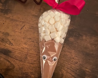 May include: A clear cellophane cone filled with brown powder and white marshmallows, decorated to resemble a reindeer. Features googly eyes, a red nose, a red ribbon, and brown pipe cleaner antlers. A festive treat.