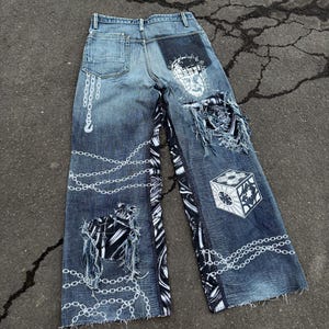 Puede incluir: Vaqueros de mezclilla desgastados con cadenas blancas y diseños gráficos. Los vaqueros tienen un gráfico en blanco y negro en la pierna izquierda y un gráfico de cubo blanco en la pierna derecha.