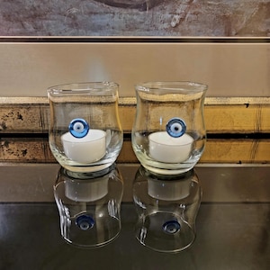 Puede incluir: Dos portavelas de vidrio transparente con un amuleto de ojo azul mal de ojo en cada uno. Cada portavelas tiene una vela de té blanca en su interior.