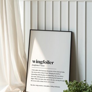 May include: A framed print with a black border displays the word "wingfoiler" in a bold font, along with its pronunciation and a definition. The definition describes a sky sailor who harnesses the power of the wind and rides the waves with a wing.