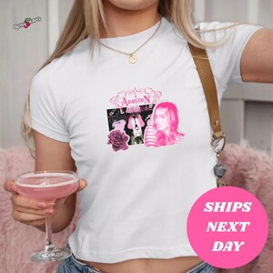 以下が含まれることがあります： ピンク色の「Addison」という名前とピンクの髪の女性の写真がプリントされた白いTシャツ。Tシャツは半袖です。「Ships Next Day」という文字が入ったピンク色の円が見えます。