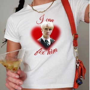 May include: White t-shirt with a red heart graphic featuring a portrait and the text "I can fix him" in red script. The shirt has short sleeves.