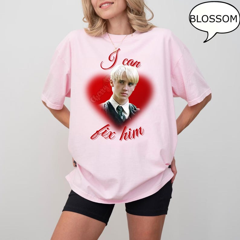 Może przedstawiać: Jasnor&oacute;żowa koszulka z grafiką w kształcie serca. Grafika zawiera wizerunek osoby i napis "I can fix him" w czerwonym skrypcie. Słowo "Blossom" znajduje się w dymku w prawym g&oacute;rnym rogu.