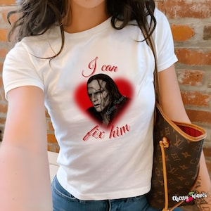 Puede incluir: Camiseta blanca con un gráfico de una persona dentro de un corazón rojo, con el texto "I can fix him" en escritura roja. La imagen tiene el texto "cherry waves" en la esquina inferior derecha.