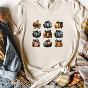 May include: A beige t-shirt with a pattern of nine blue and orange pumpkins with black bows. The pumpkins are arranged in a grid pattern.