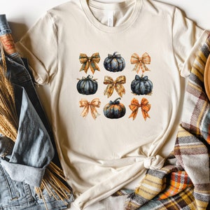 May include: Cream-colored t-shirt featuring a grid of watercolor pumpkins and bows in shades of orange, navy, and gold. The shirt is styled with a denim jacket, a plaid scarf, and a brown hat, creating a fall-themed aesthetic.