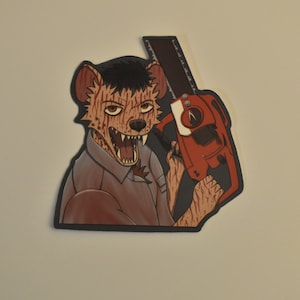May include: A sticker featuring a cartoon hyena with a chainsaw. The hyena is wearing a brown shirt and has a fierce expression. The chainsaw is red and black.