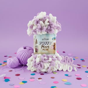 May include: A still-life image of Alize Puffy More Plus yarn in a can, with a ball of purple yarn and a small ball of purple yarn. The yarn is white and lavender. The background is a solid purple with colorful confetti.
