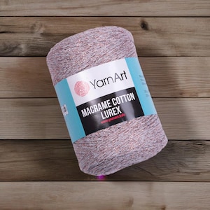 Può includere: Un gomitolo di filato YarnArt Macrame Cotton Lurex. Il filato è una miscela di colori viola chiaro e rame. L'etichetta è azzurra e mostra il logo YarnArt e le informazioni sul prodotto.